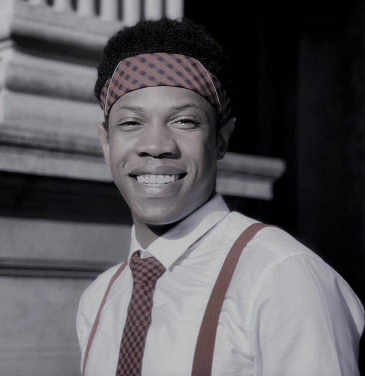 A black man with a broad welcoming smile. He wears a shirt with a tie and suspenders with a head scarf around his forehead.