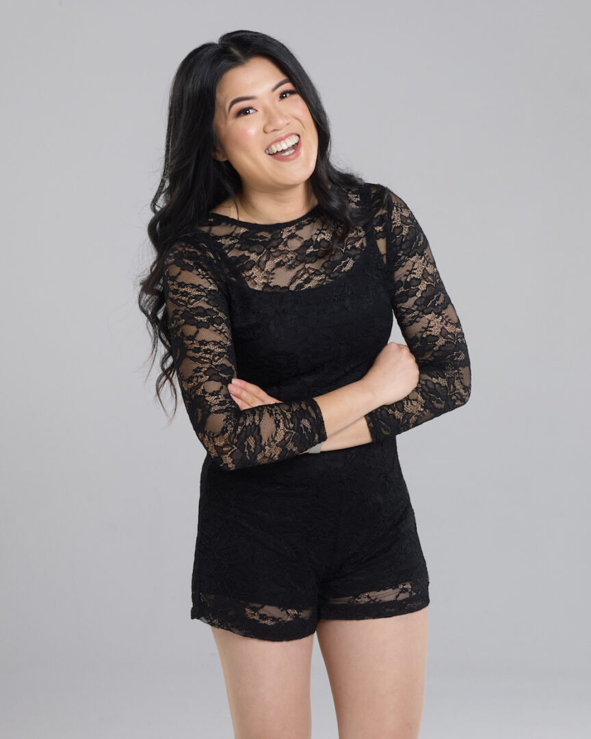 a woman stands in front of a grey background, she is smiling and has her arms crossed in front of her. She is wearing a black lace jumpsuit, her hair is curled and falls around her
shoulders.