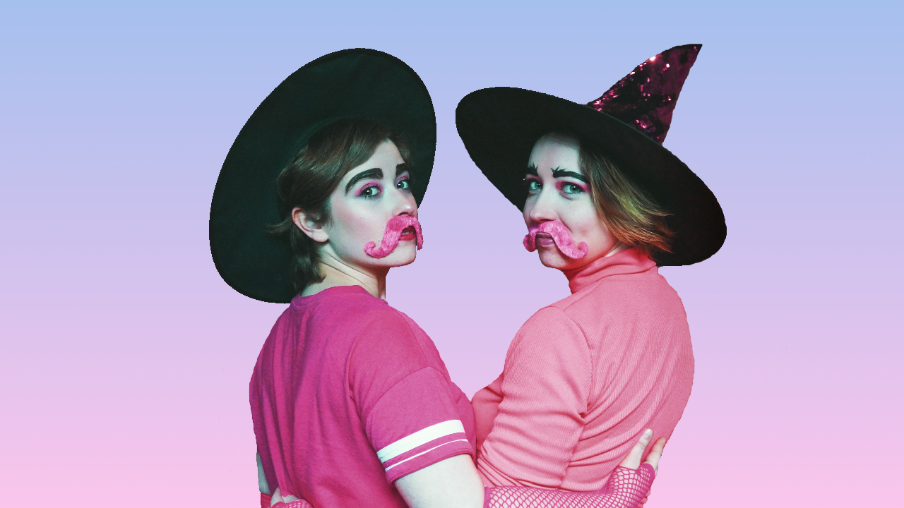 Two non-binary people stand in front of a pink background, looking over their shoulders. They are wearing pink shirts, pink moustaches, and witches hats