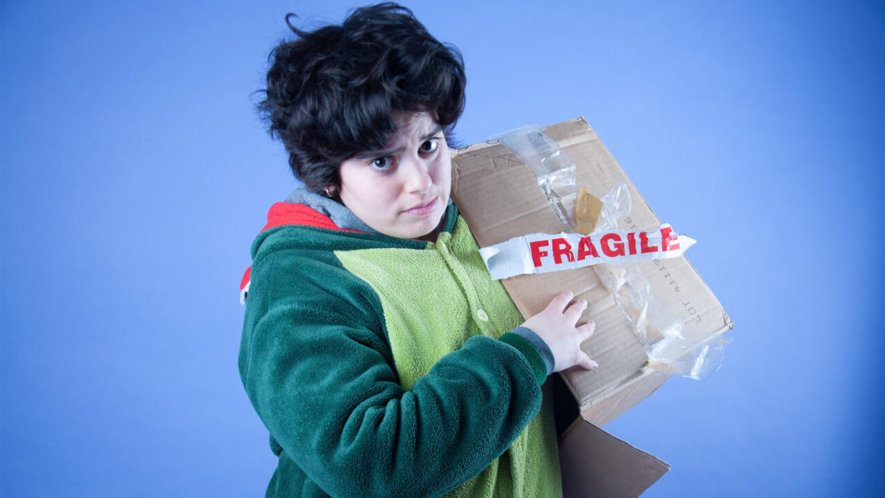 Artemis looks directly at the camera with an expression of concern. They have olive toned skin and short, curly, dark brown hair. They are wearing a green dinosaur onesie. One of their arms is deep within a cardboard box which is labeled ‘fragile’. The background of the image is a pale blue colour.