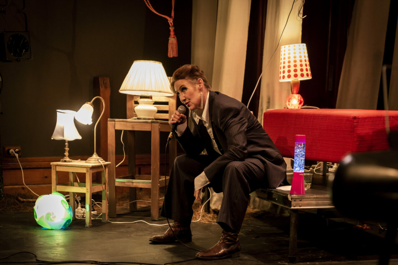 A woman in a suit sits on a bench speaking into a microphone, she is surrounded by lamps of various shapes and sizes.