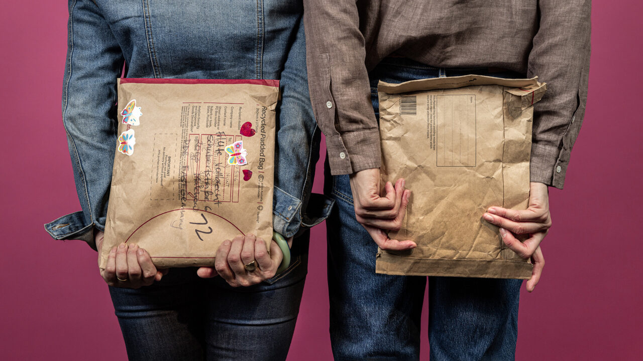 Parcels are held behind two women’s backs, one is wearing a denim jacket, the other a brown shirt, against a pink background.