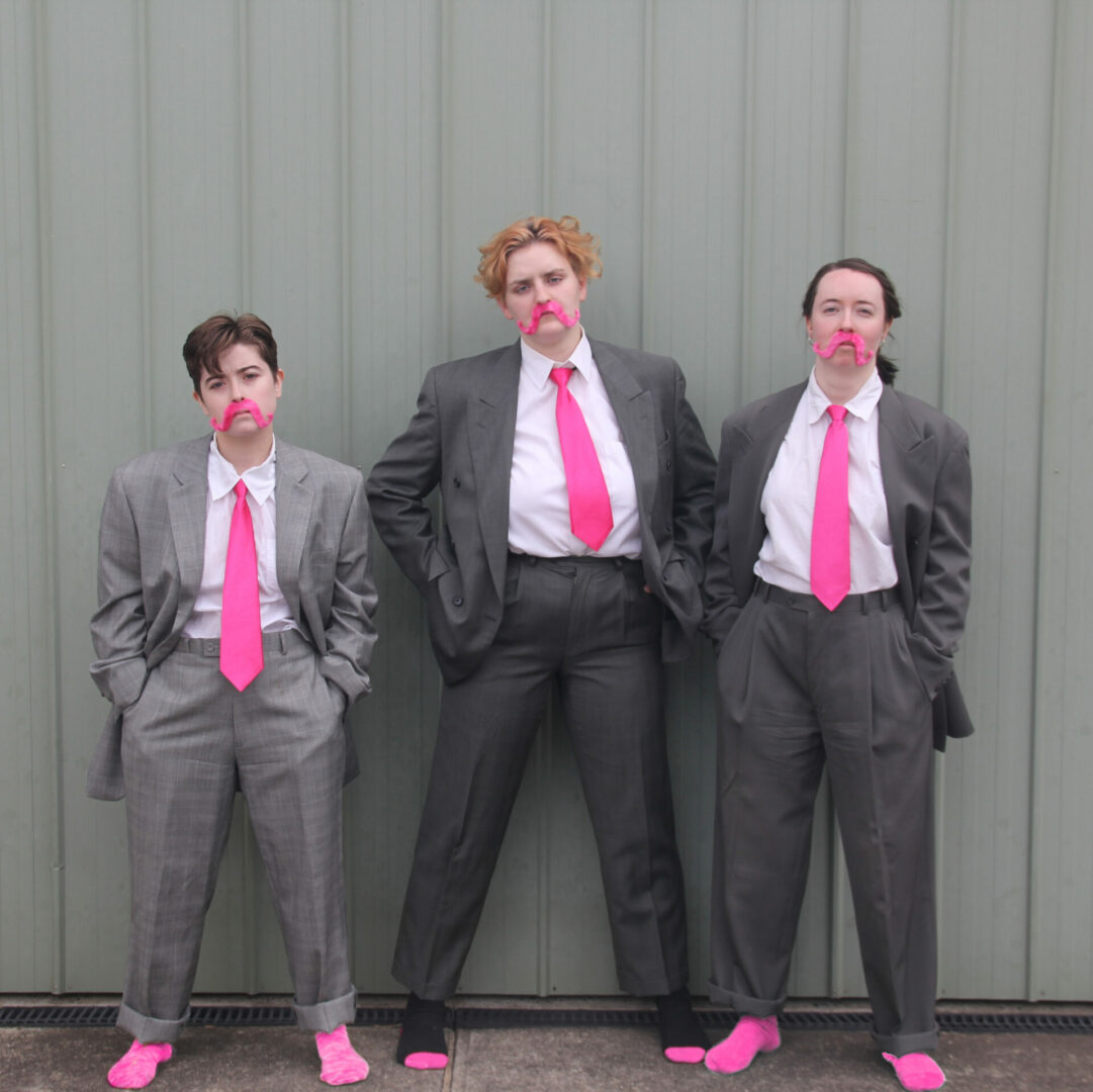 Three men wearing suits, with pink ties and pink moustaches.