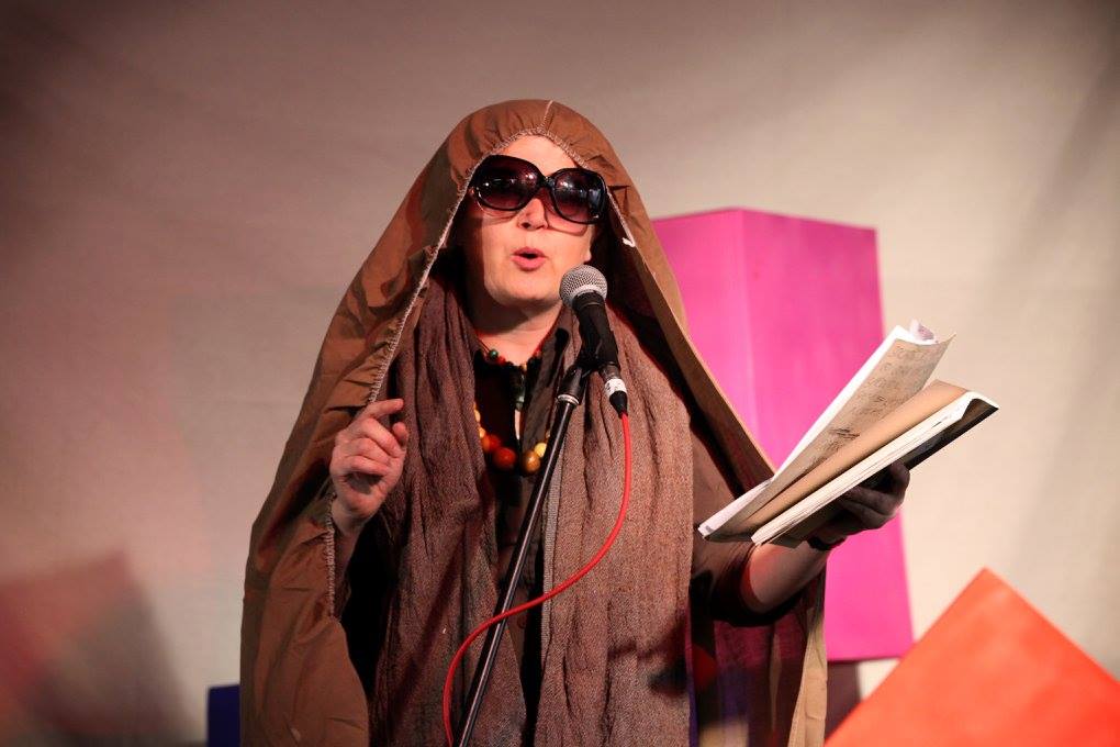 A woman stands inside a brown sheet, she holds papers in her left hand and wears sunglasses.