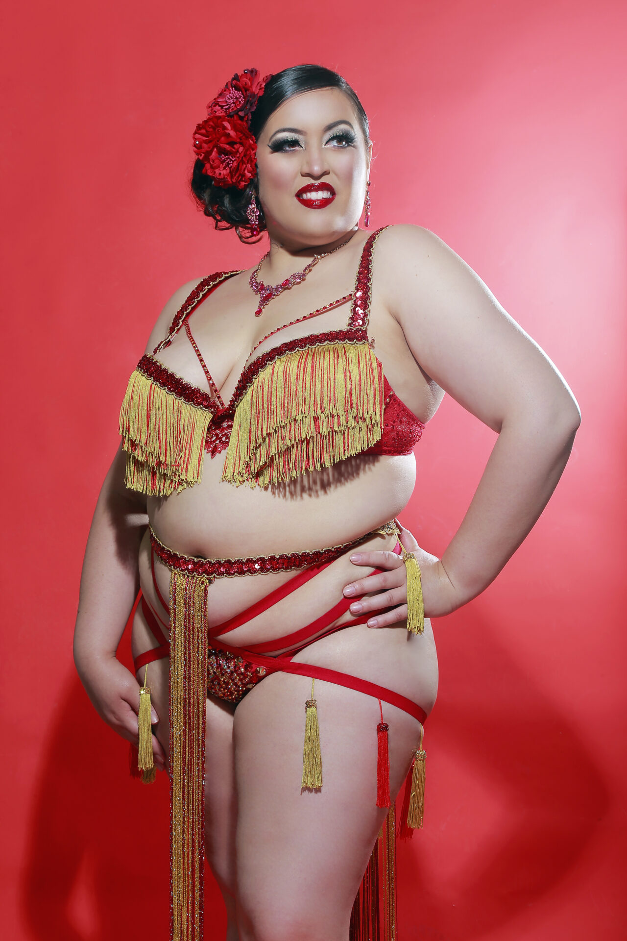 A woman in a red and gold fringed bra and g-string, wearing a long fringed red and gold skirt on a red background with brown hair in an updo pinned by a red rose