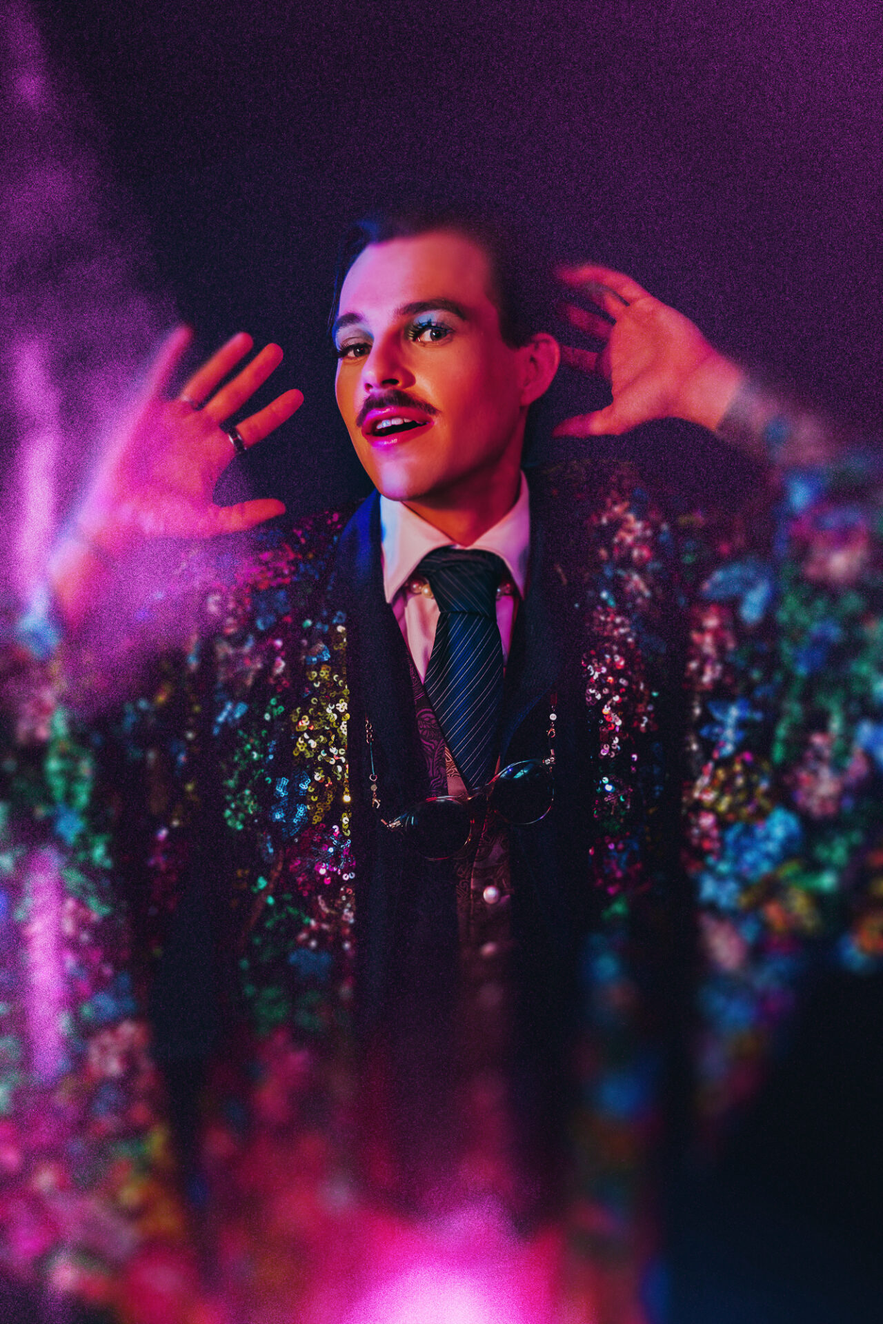 A moustachioed person in a colourful three-piece suit and tie, with a rainbow sequinned kimono gazes at the camera