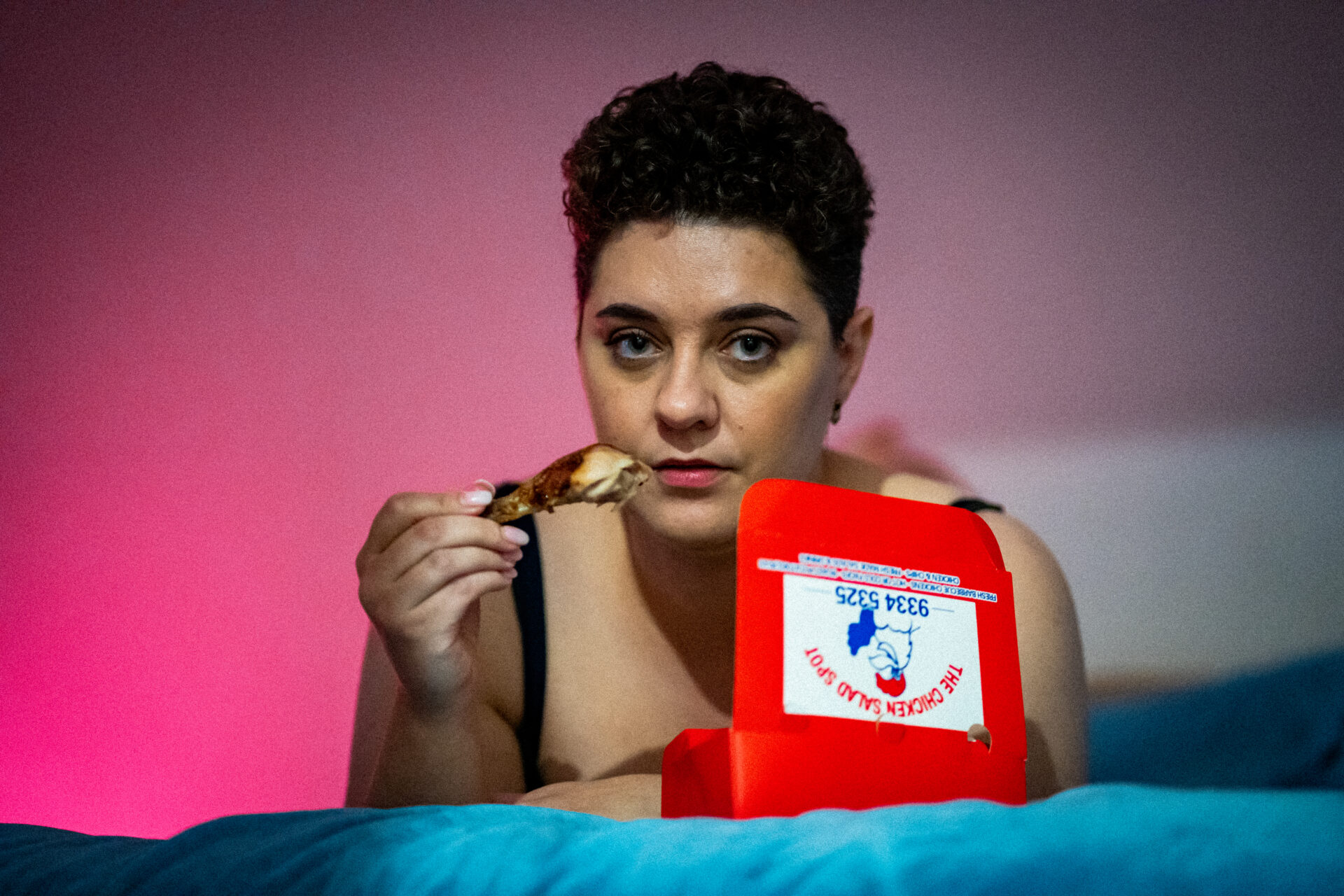 A woman lying on a bed, seductively holding a roast chicken drum stick out of a red takeaway box. Pink lighting in background 