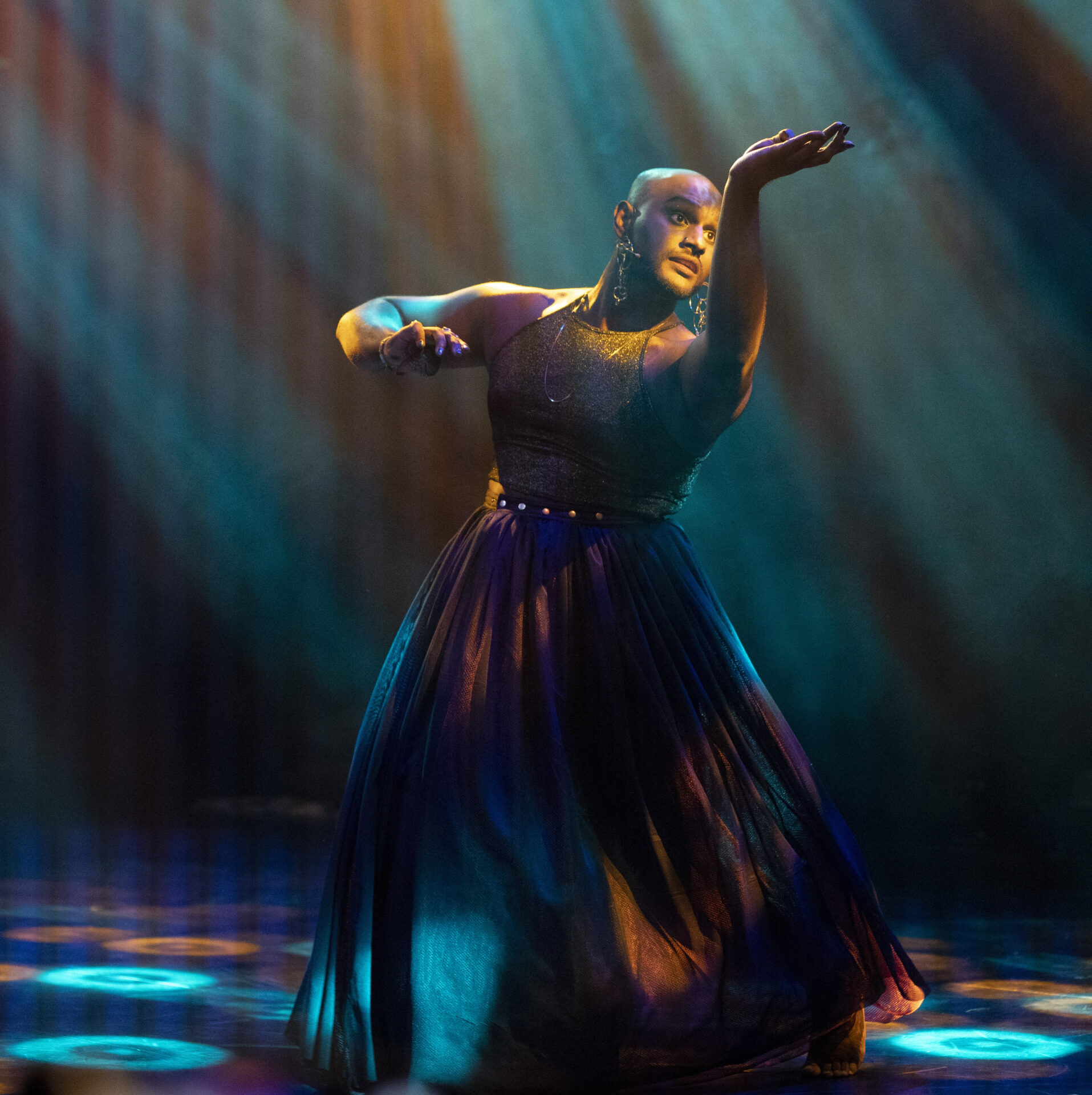 A brown person wearing a purple skirt and a golden top is moving against a backdrop of shiny, dark lighting.