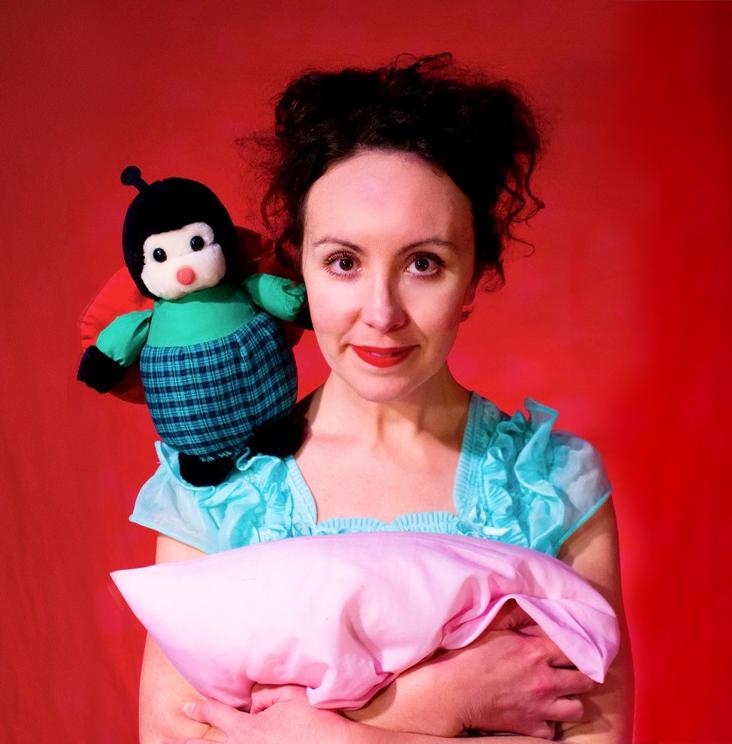 A woman with dark hair and brown eyes is looking at the camera with a whimsical expression. She is wearing a mint green nightie and cuddling a light pink pillow to her chest. A cute little bug toy is resting on her shoulder. 